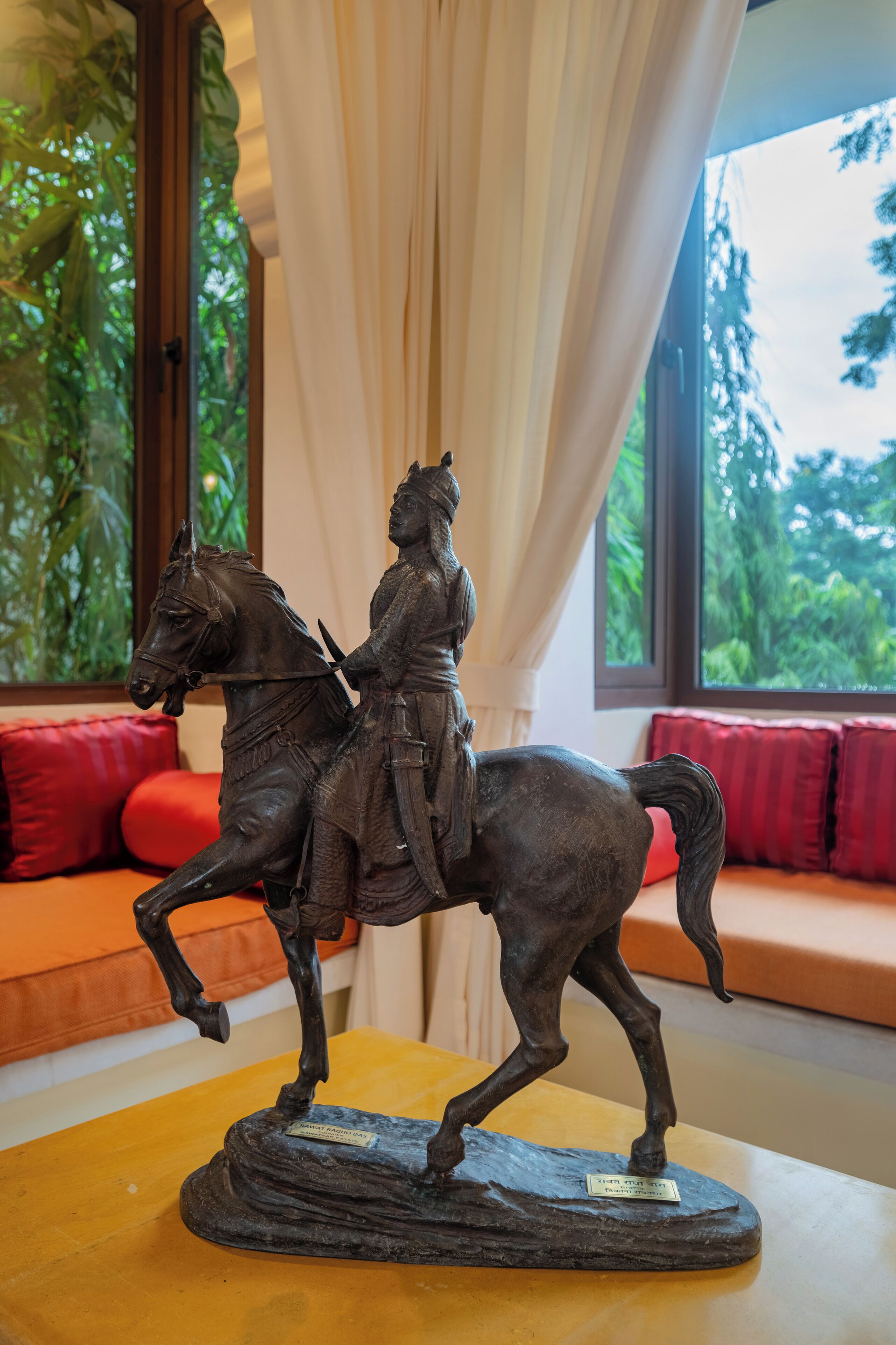 Una estatua de una figura a caballo sobre una mesa en una habitación con ventana, cojines rojos y una planta.