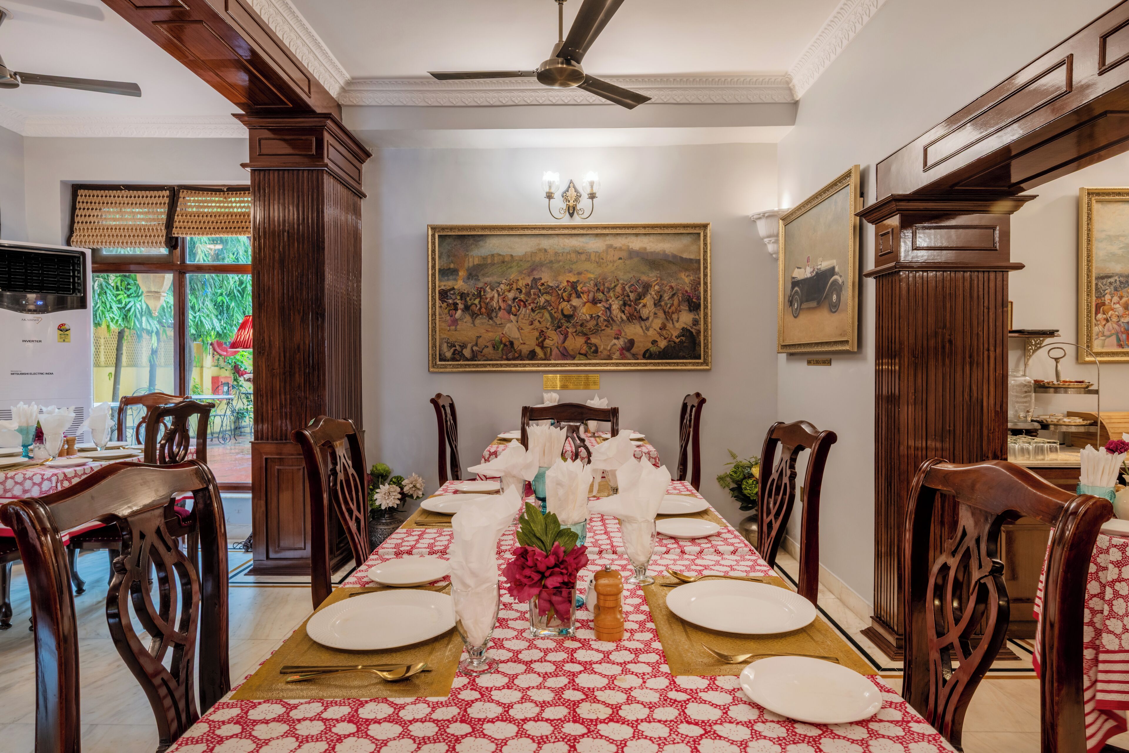 A dining area with a long table set for a meal, wooden chairs, a ceiling fan, and a large painting on the wall.