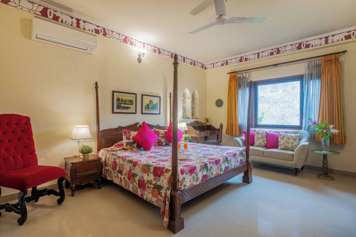 A bedroom with a four-poster bed, a red armchair, a sofa, a small table, and a window with curtains.