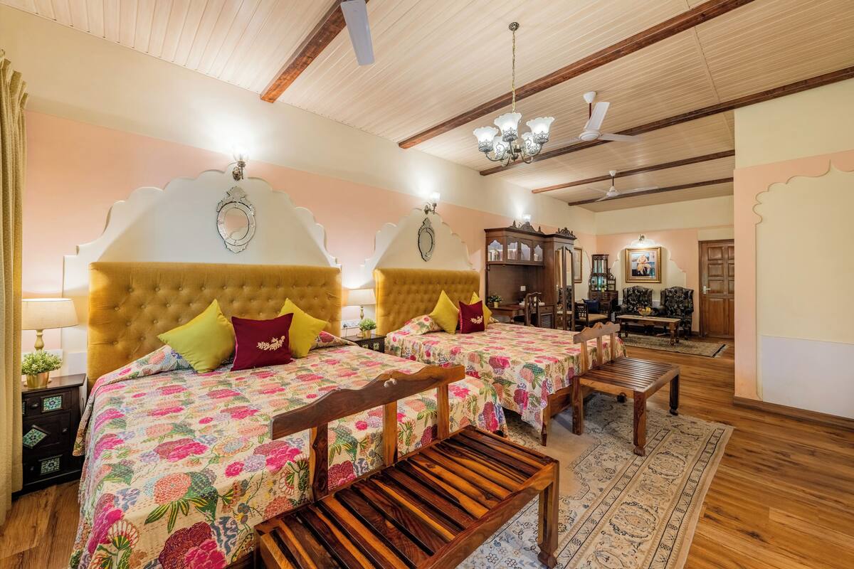 A hotel room with two beds, wooden headboards, floral bedding, a wooden bench, a chandelier, and a ceiling fan.