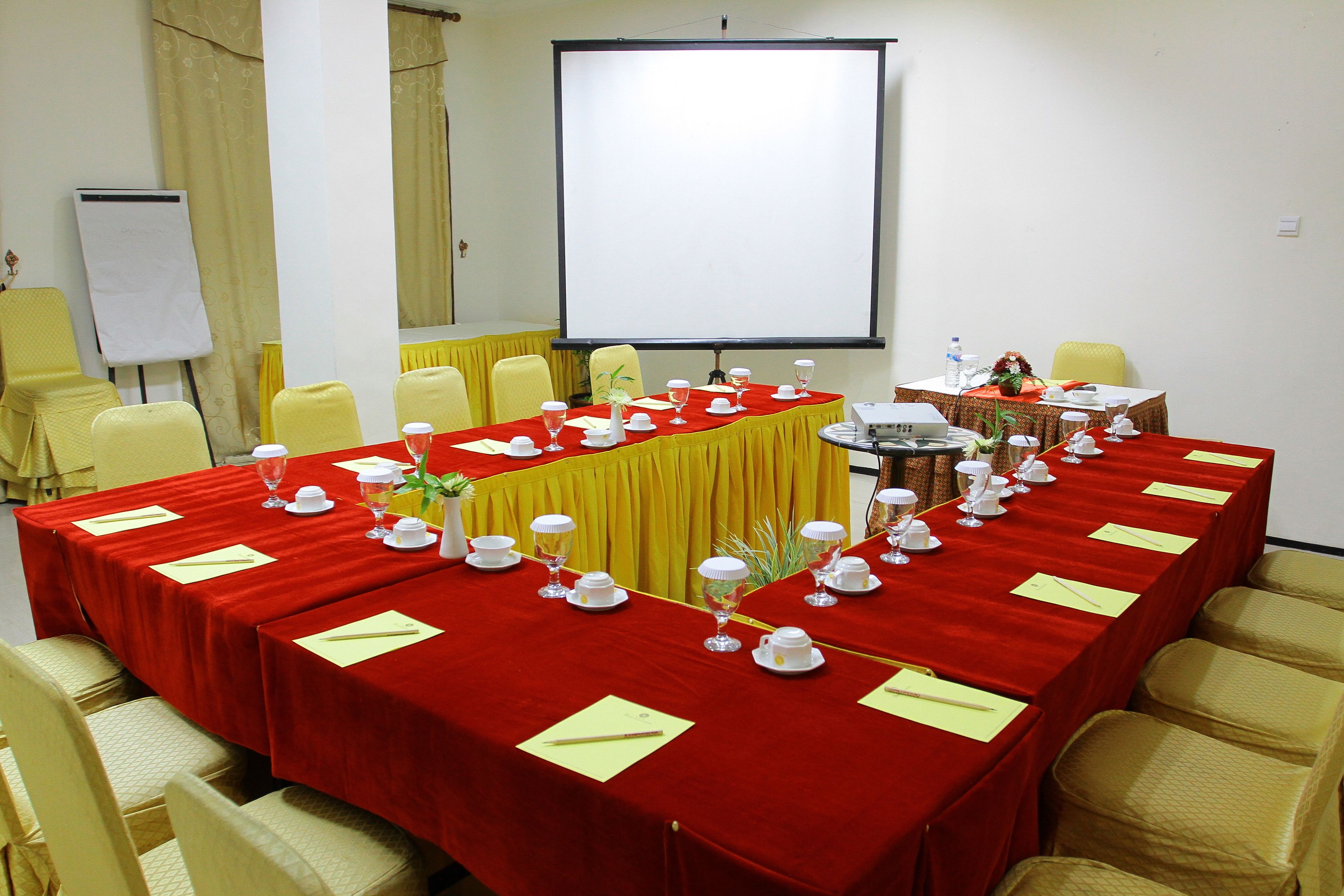 Un salón de conferencias con una larga mesa dispuesta para una reunión, sillas amarillas, una pizarra y una pantalla de proyector.