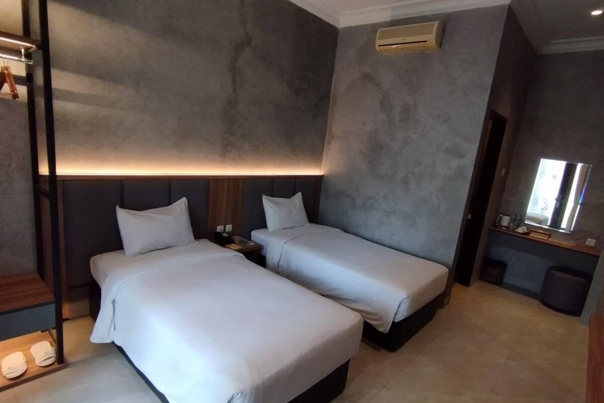 Ein Hotelzimmer mit zwei Einzelbetten, einem Nachttisch mit Lampe, einem Spiegel und einer kleinen Sitzecke mit Stuhl und Tisch.