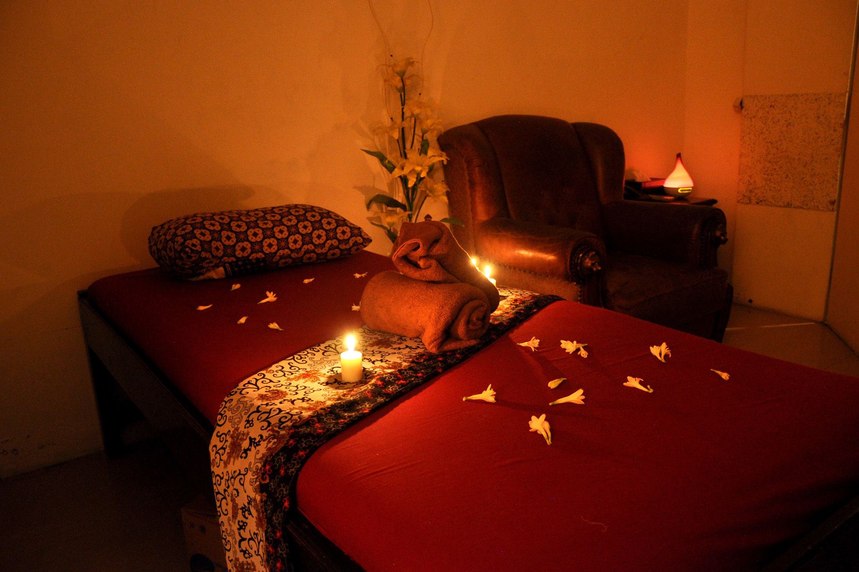 A room with two beds, a chair, a vase with flowers, and a lit candle.