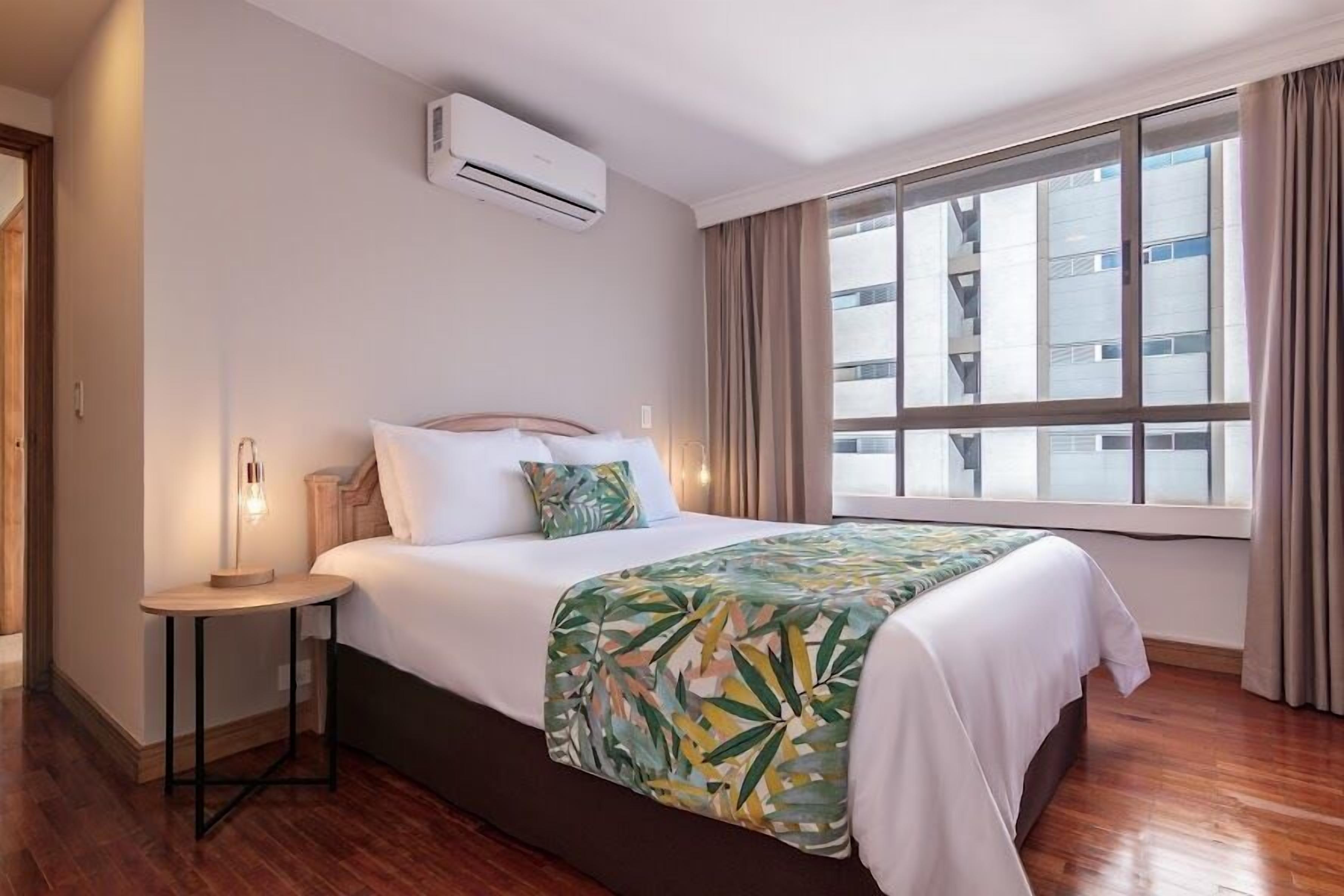 Habitación de hotel con cama, mesita de noche, equipo de aire acondicionado, ventana con cortinas y vista a un edificio.