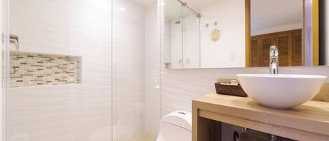 Un baño moderno con cabina de ducha de vidrio, inodoro blanco y tocador de madera con un lavamanos grande y blanco.