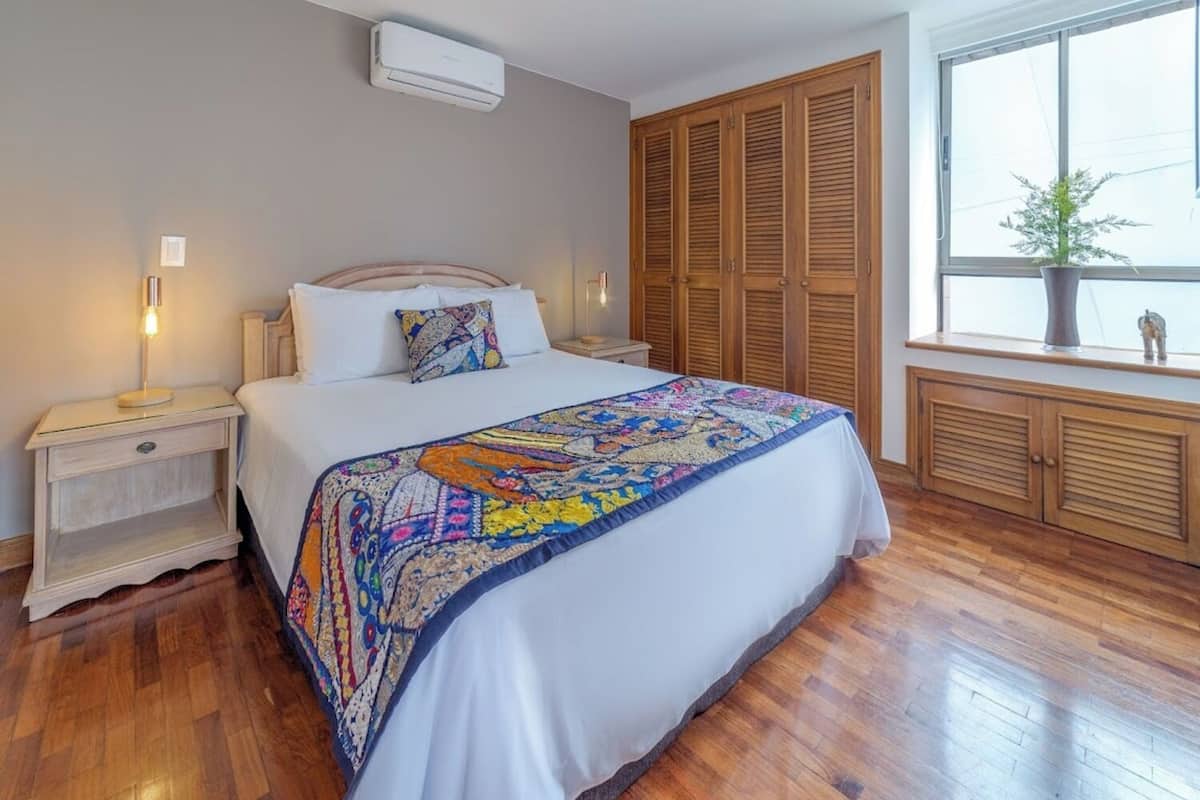 Une chambre à coucher avec un lit, une armoire en bois, une table de chevet, une lampe et une fenêtre agrémentée d’une plante.