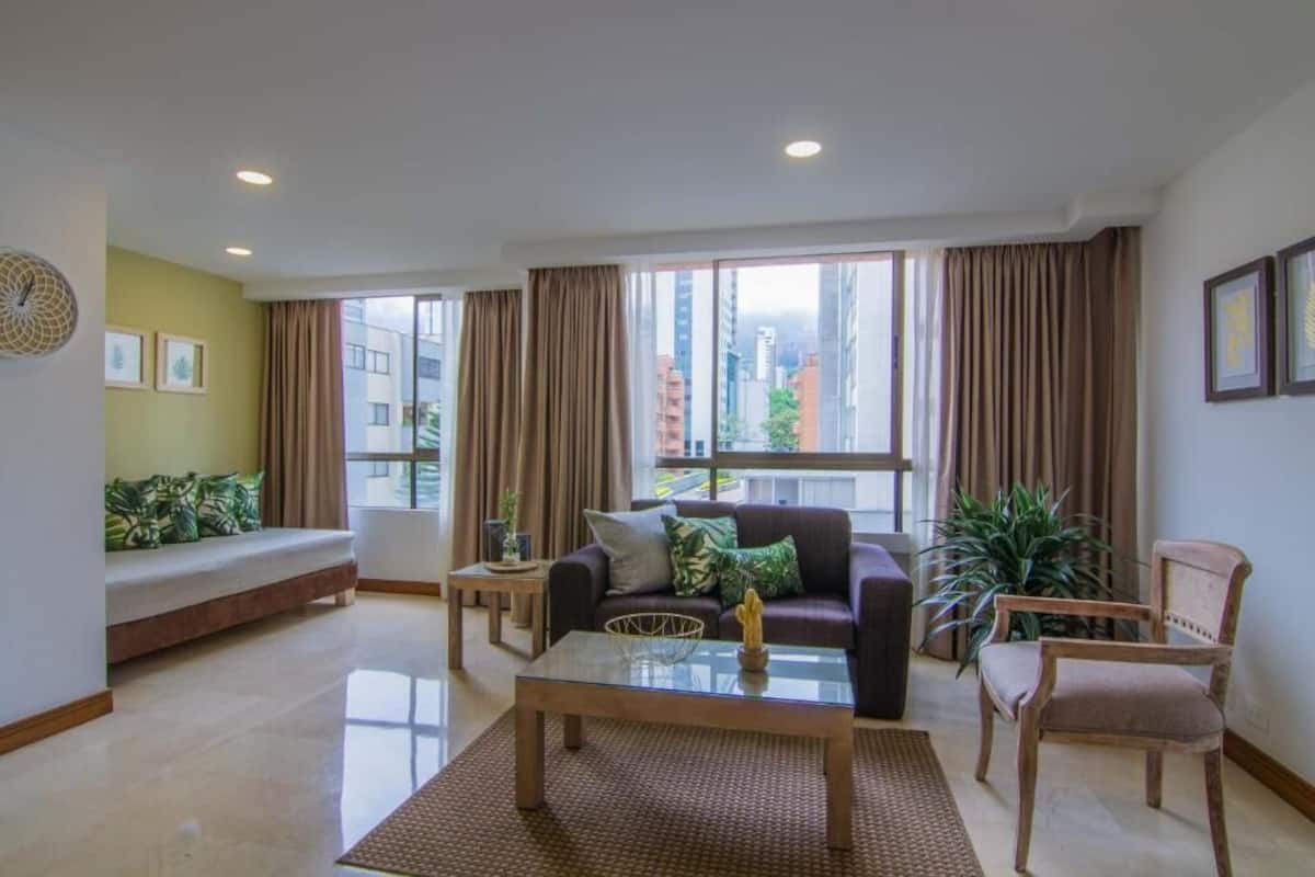 Un salon moderne avec un canapé, une table basse et des fauteuils, de grandes fenêtres avec des rideaux et une vue sur les bâtiments environnants.
