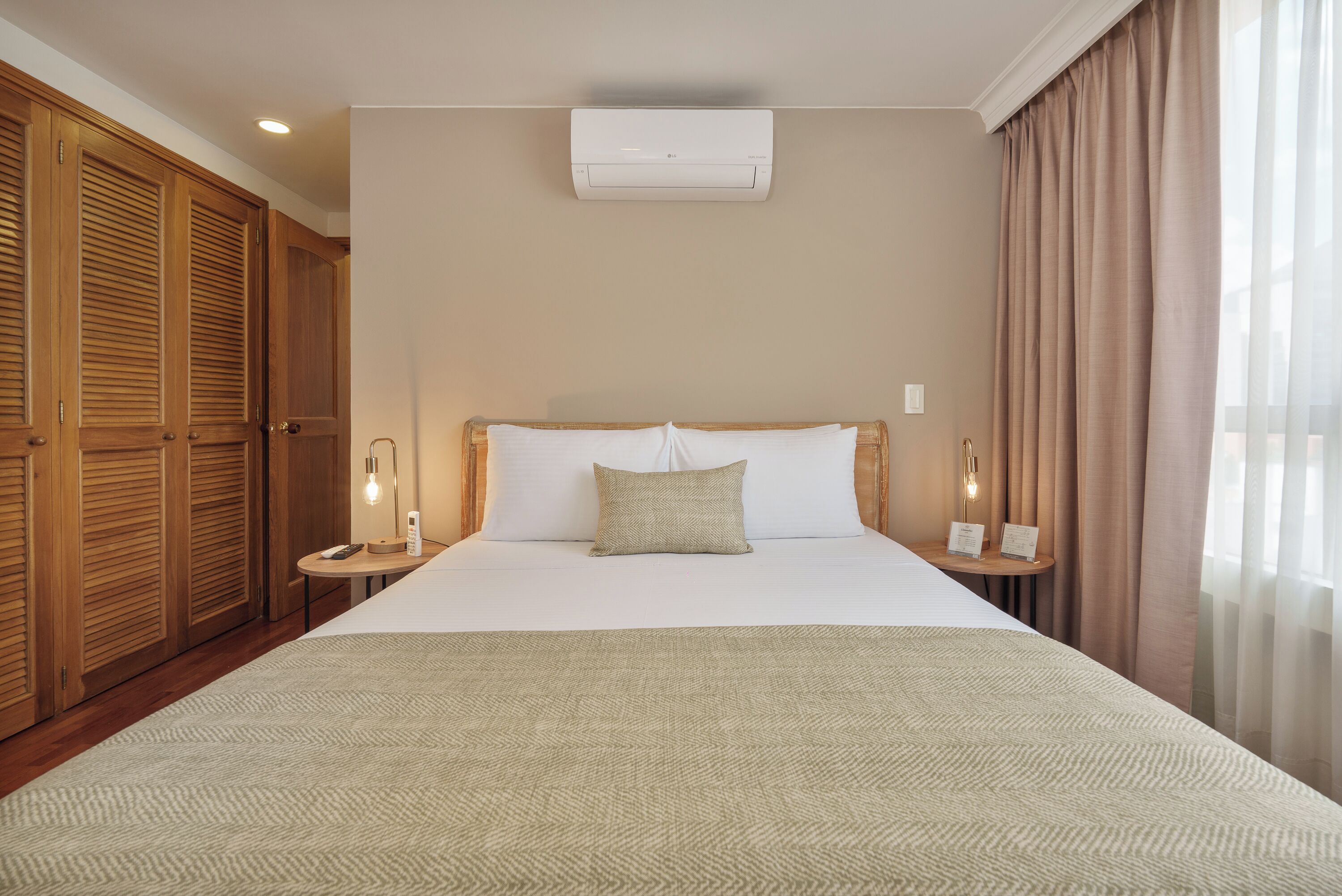 A hotel room with a large bed, wooden wardrobe, bedside lamps, and an air conditioning unit.