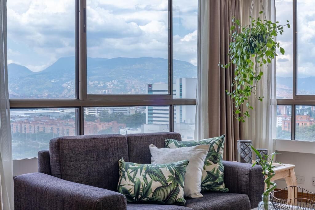 Ein modernes Wohnzimmer mit grauem Sofa, Kissen mit tropischem Muster, einer hängenden Pflanze und Blick auf eine Stadtlandschaft durch große Fenster.