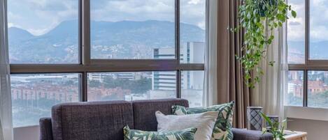 Ein modernes Wohnzimmer mit grauem Sofa, Kissen mit tropischem Muster, einer hängenden Pflanze und Blick auf eine Stadtlandschaft durch große Fenster.
