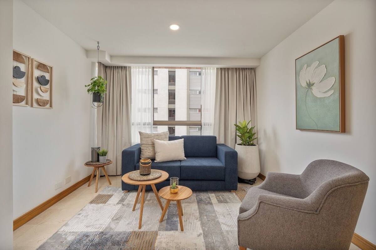 Un salon moderne avec un canapé bleu, un fauteuil gris et un tapis à motifs. On y trouve deux petites tables en bois ornées d’objets décoratifs. Une grande fenêtre avec des rideaux est visible, et un tableau est accroché au mur.