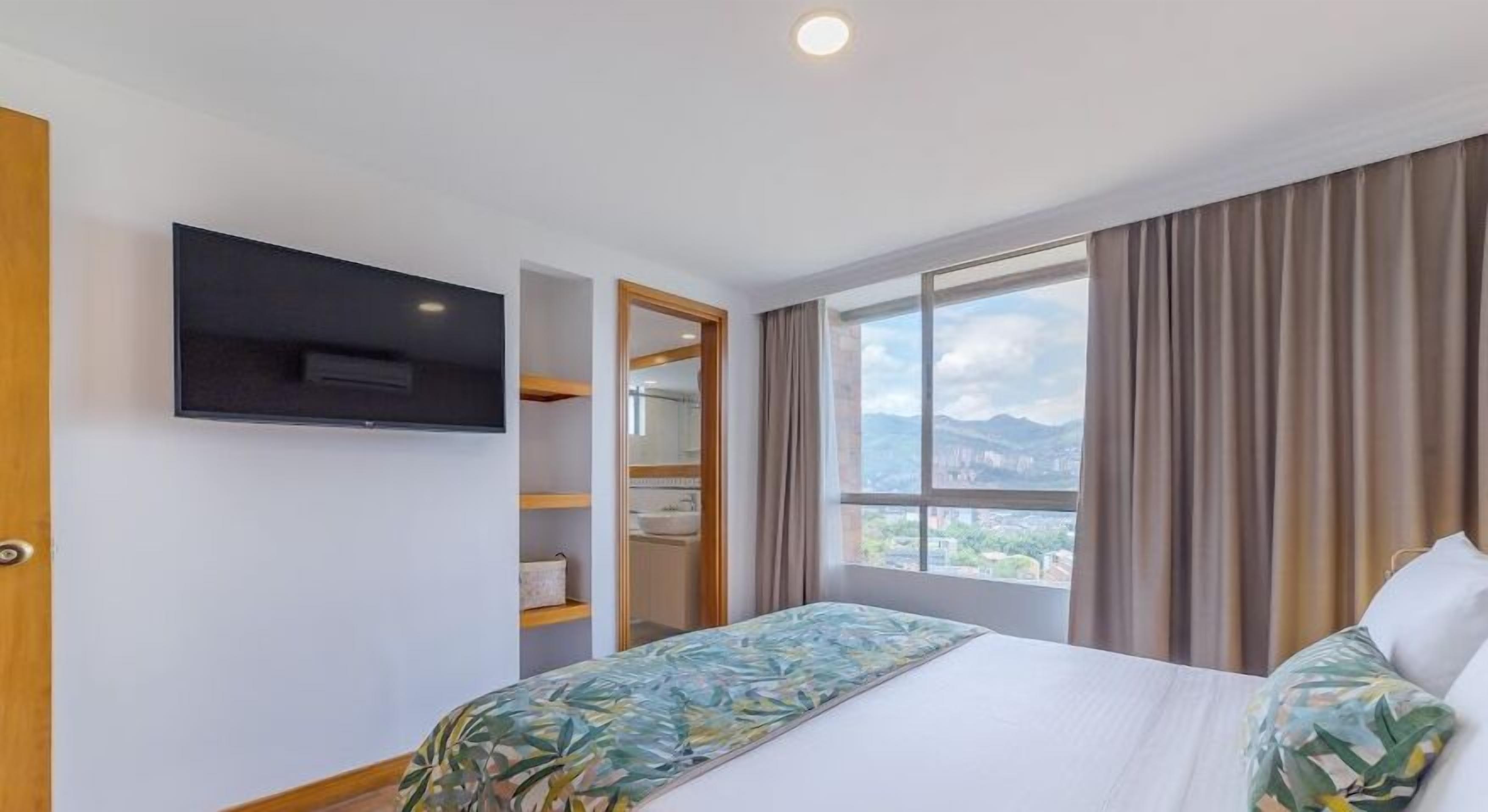 Habitación de hotel con cama, televisor montado en la pared, baño con espejo y lavamanos, y ventana con vistas a las montañas.