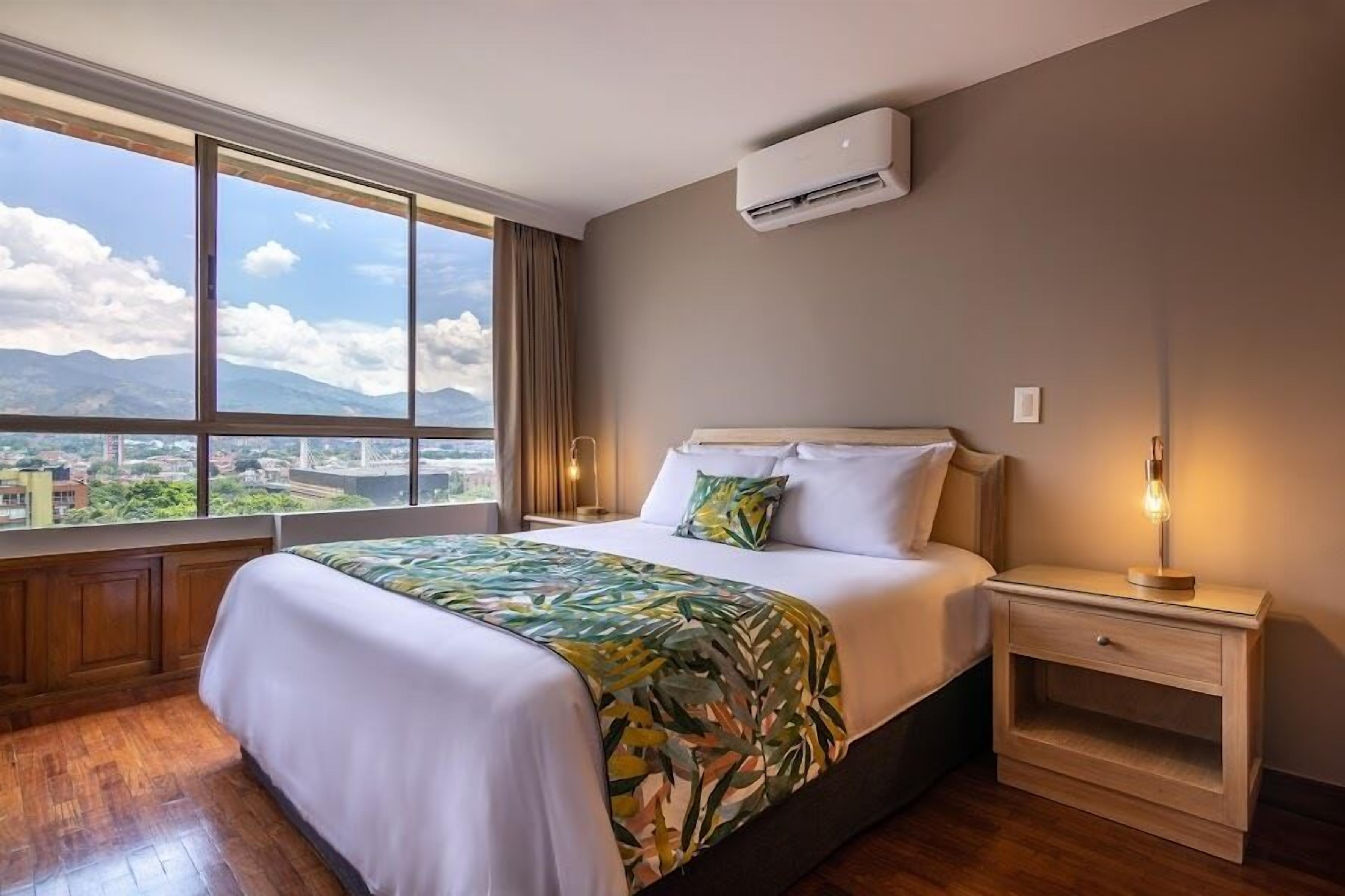 A hotel room with a large window, a bed with a floral bedspread, a nightstand with a lamp, and an air conditioning unit on the wall.