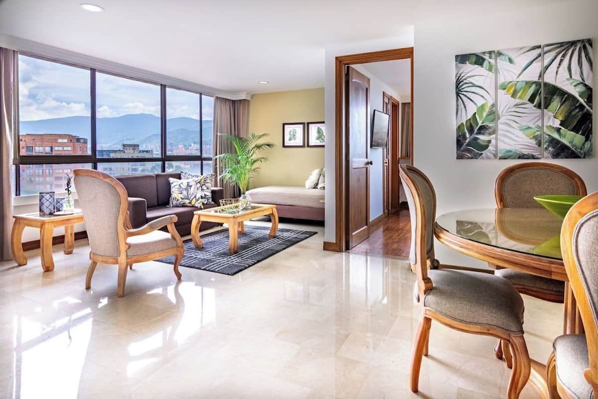 Un salon moderne comprenant un canapé, des fauteuils, une table basse et une table à manger. Une grande fenêtre offre une vue sur les bâtiments et les montagnes.