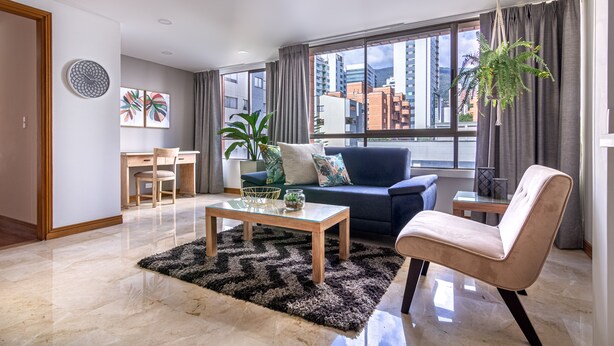 Una sala de estar moderna con una mesa de centro de vidrio, un sofá, un sillón y un ventanal con cortinas.