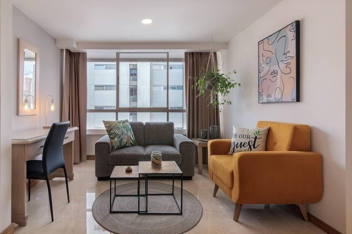 Un salon moderne avec un canapé gris, un fauteuil jaune, une table basse et un bureau avec une chaise. On y trouve une grande fenêtre avec des rideaux, une plante et une œuvre d’art murale.