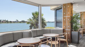 A modern interior with a large window overlooking a body of water, a seating area with a sofa and round tables, and a wooden accent wall. - Lakes Hotel (West Lakes)