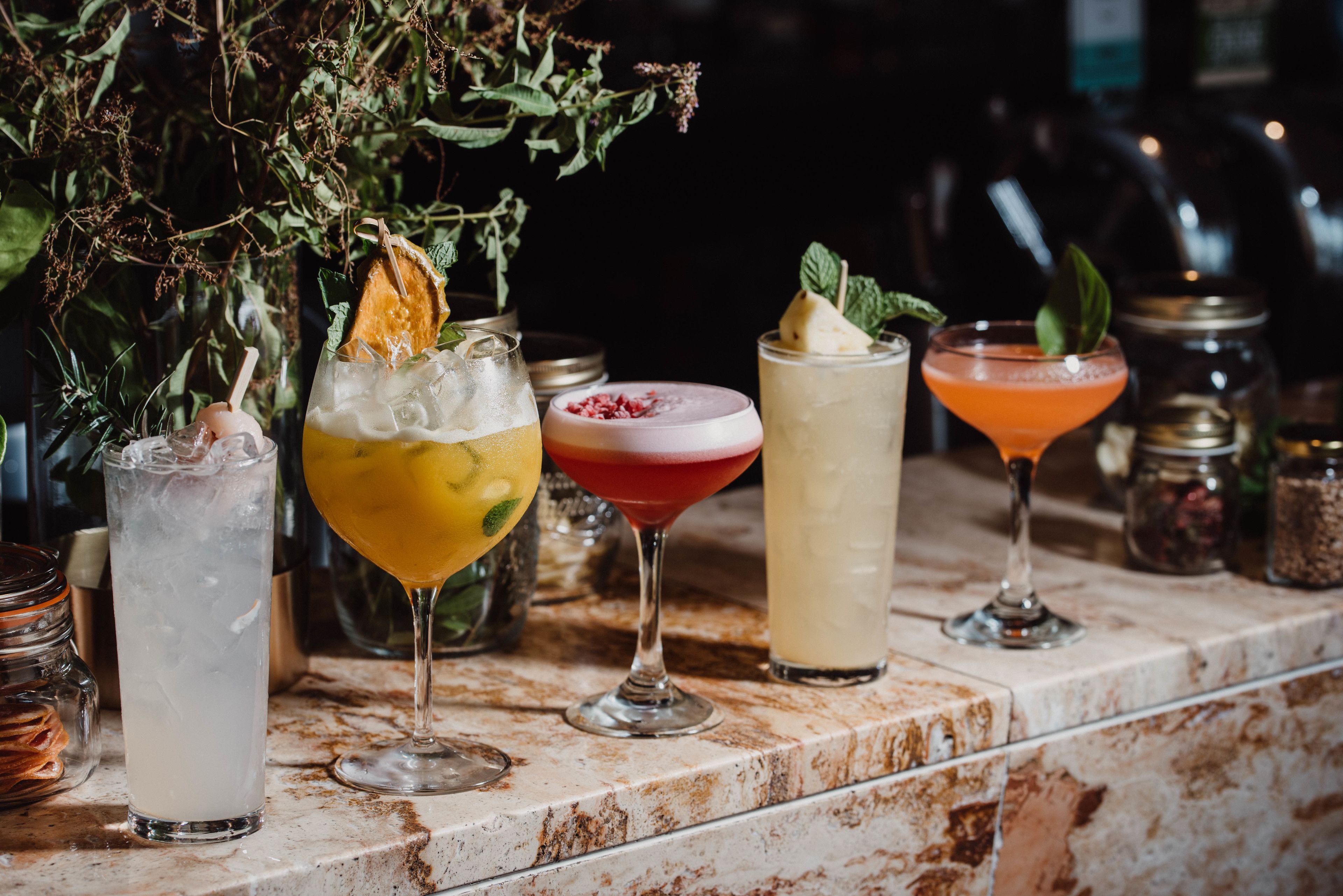 A bar counter with various drinks, including a tall glass with ice, a yellow drink with a lemon slice, a red drink with a garnish, a clear drink with a mint leaf, and an orange drink in a glass.