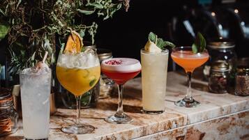 A bar counter with various drinks, including a tall glass with ice, a yellow drink with a lemon slice, a red drink with a garnish, a clear drink with a mint leaf, and an orange drink in a glass.
