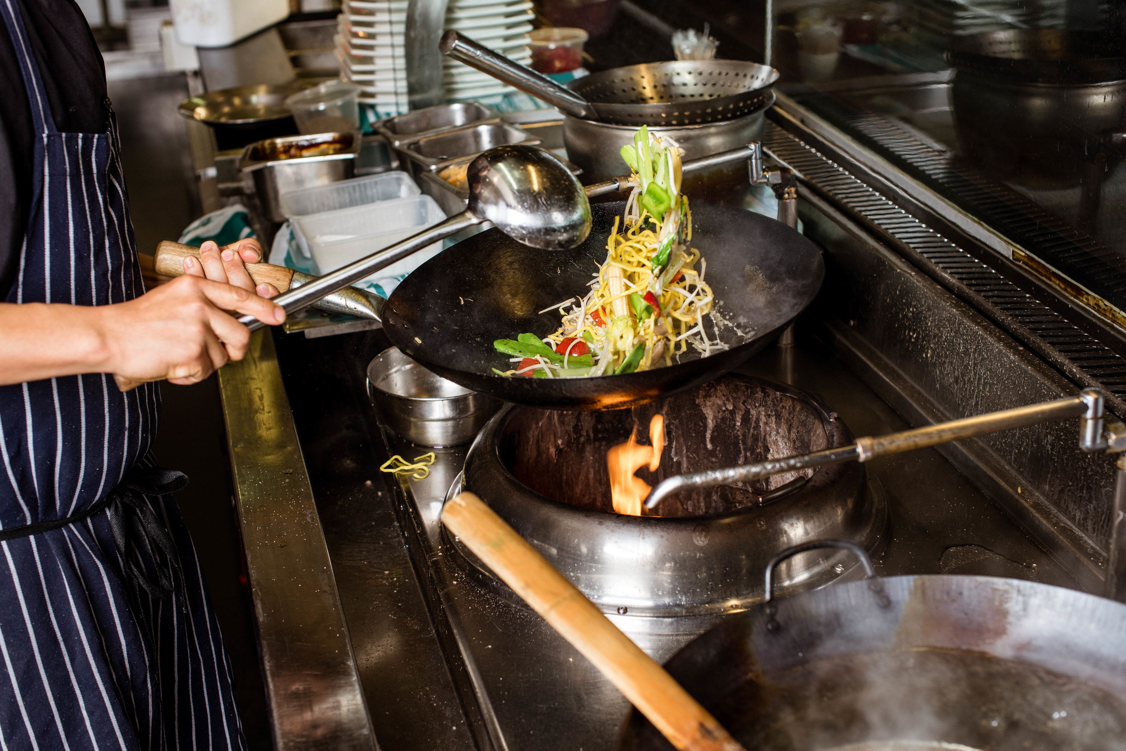 A kitchen with a wok on a stove, a ladle, and a person cooking.