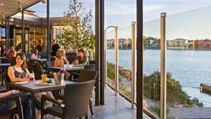 An outdoor dining area with tables and chairs, overlooking a body of water. - Lakes Hotel (West Lakes)