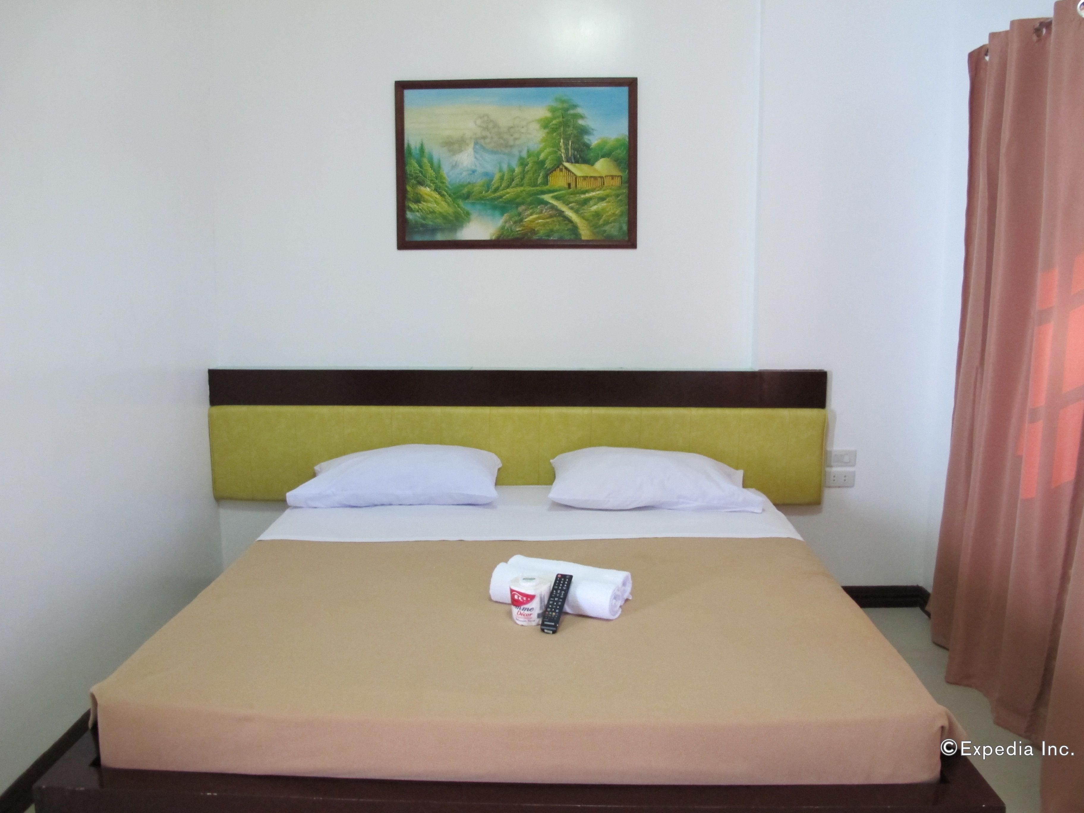 A hotel room with a bed, pillows, a remote control, and a painting on the wall.