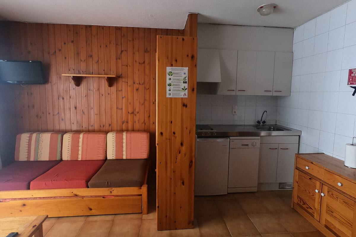 Habitación pequeña con cama de madera, kitchenette y un pequeño televisor montado en la pared.