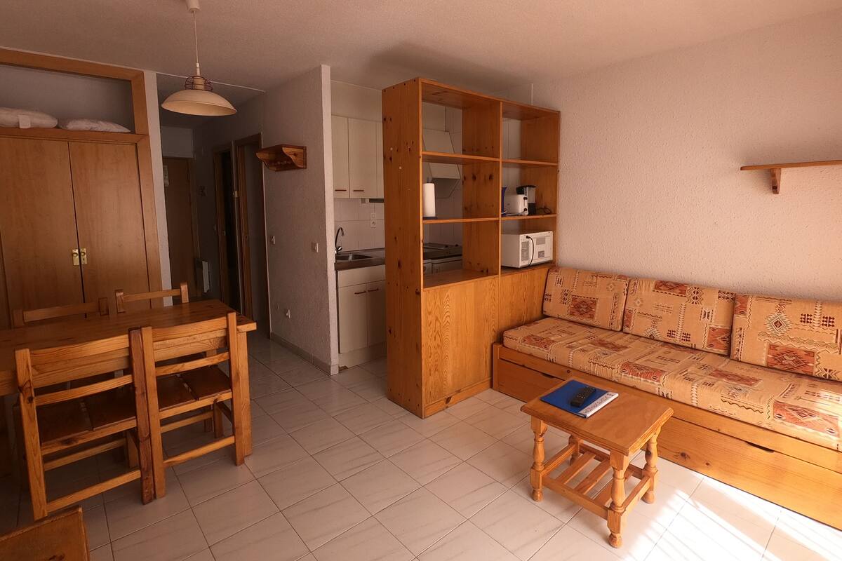 Un espacio habitable compacto con una mesa y sillas de madera, un sofá con cojines estampados, un armario de madera y una zona de kitchenette.