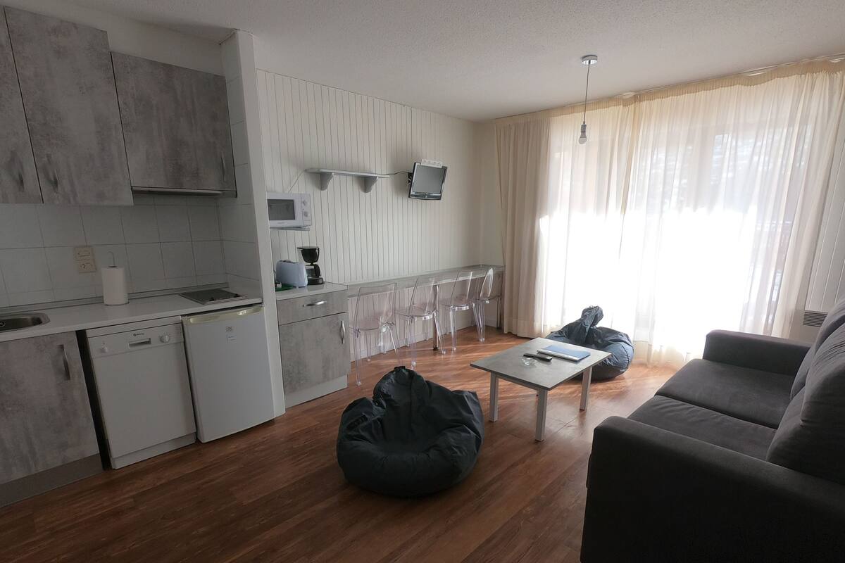 Una cocina moderna con armarios blancos, un microondas y una cafetera. Hay una zona de comedor con mesa y sillas, y un sillón de tumba en la sala de estar.