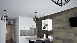 Une cuisine moderne avec des armoires blanches, un comptoir en granit et un four à micro-ondes encastré. On y trouve deux chaises noires avec des pieds en bois et une table noire. La pièce est dotée d’un plancher en bois et d’un mur recouvert de panneaux en bois foncé.
