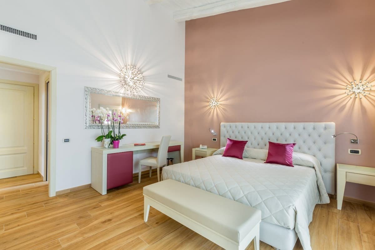 A bedroom with a large bed, a bedside table, a desk with a chair, a bench, and a mirror on the wall.