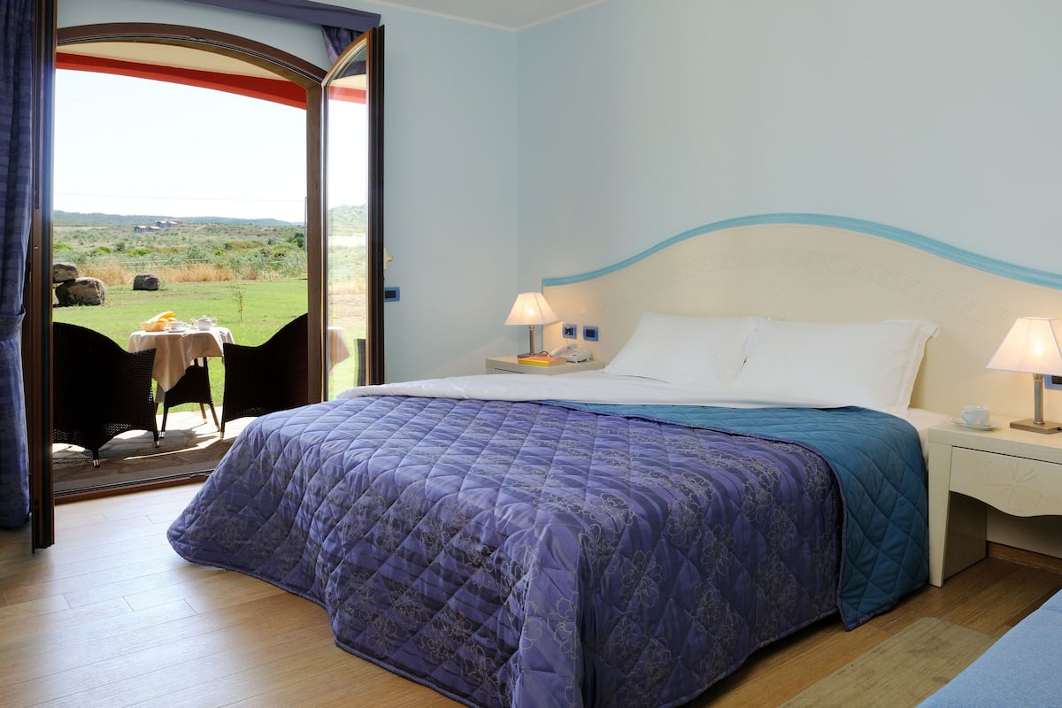 A bedroom with a bed, two bedside tables, a window with a view of the outdoors, and a door leading to a balcony.