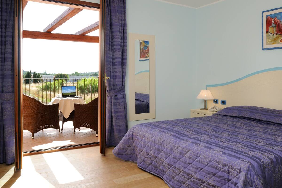 A bedroom with a bed, a nightstand, a lamp, a painting, and a view of a vineyard through an open door.