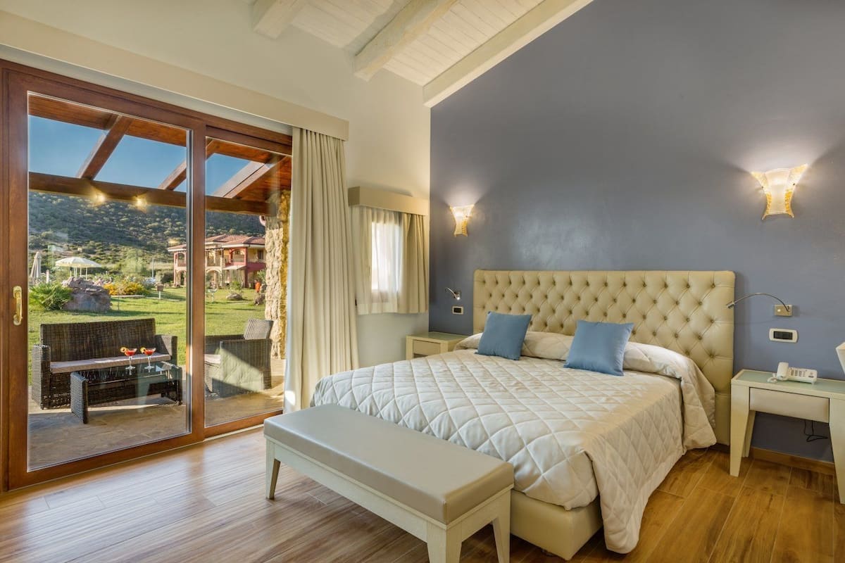 A bedroom with a large bed, a bench, a nightstand, and a view of a garden through a sliding glass door.