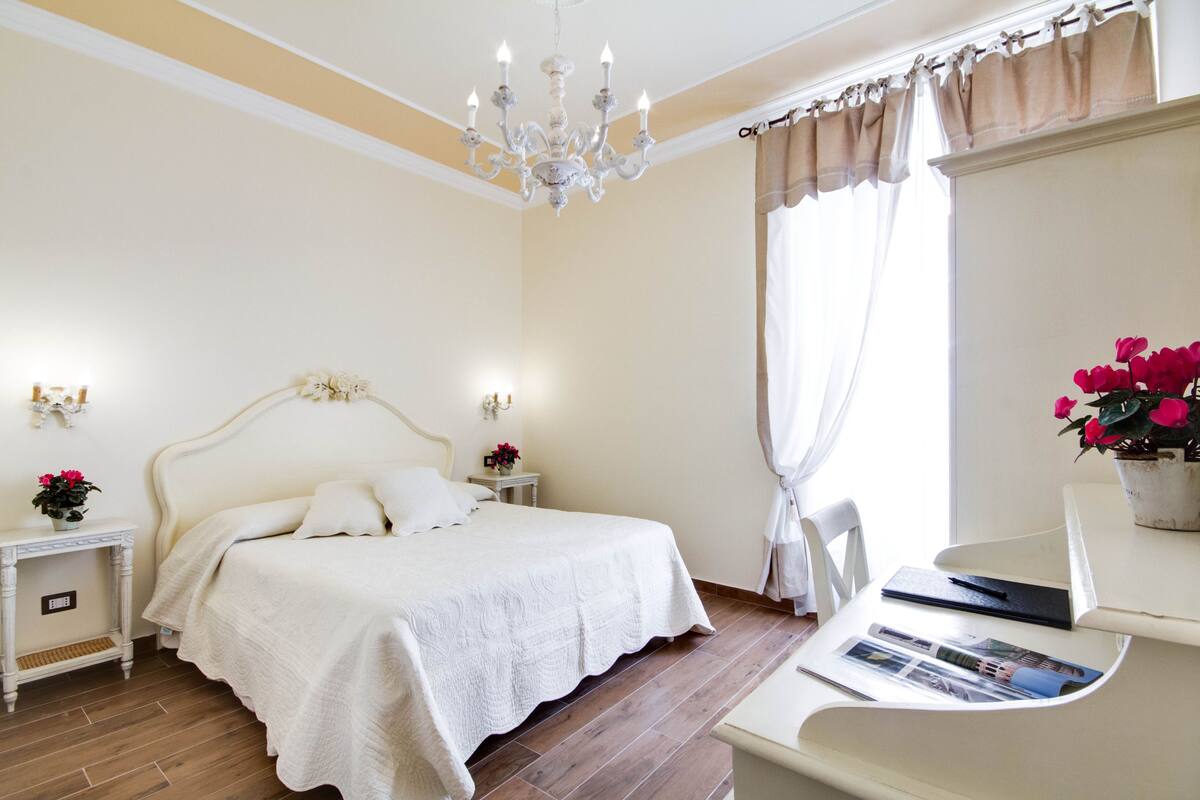 Una camera da letto con un letto grande, comodini, una scrivania con una sedia, un lampadario e una finestra con tende.