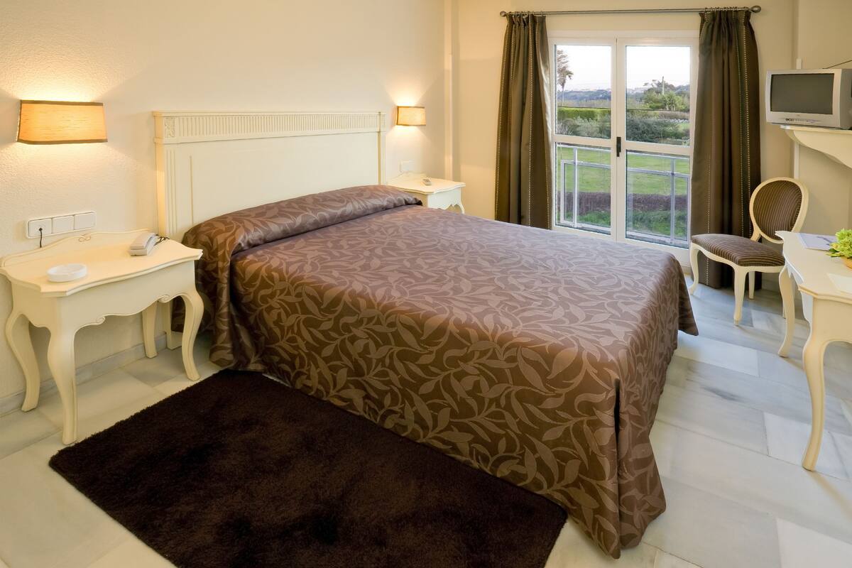 Quarto com cama, mesas de cabeceira, cadeira, escrivaninha, televisão e vista para o exterior.