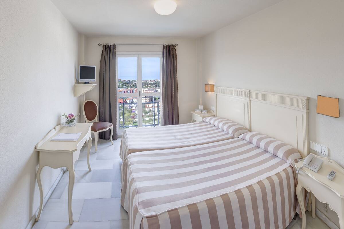 Quarto de hotel com duas camas, uma mesa, uma cadeira, uma televisão e uma janela com cortinas.