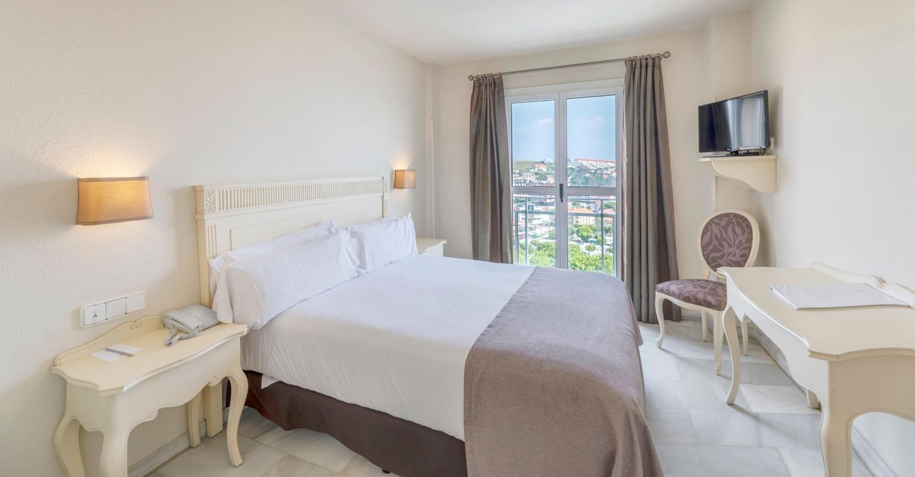 Una habitación de hotel con una cama, una mesita de noche con lámpara, un escritorio con silla, un televisor y una ventana con cortinas.