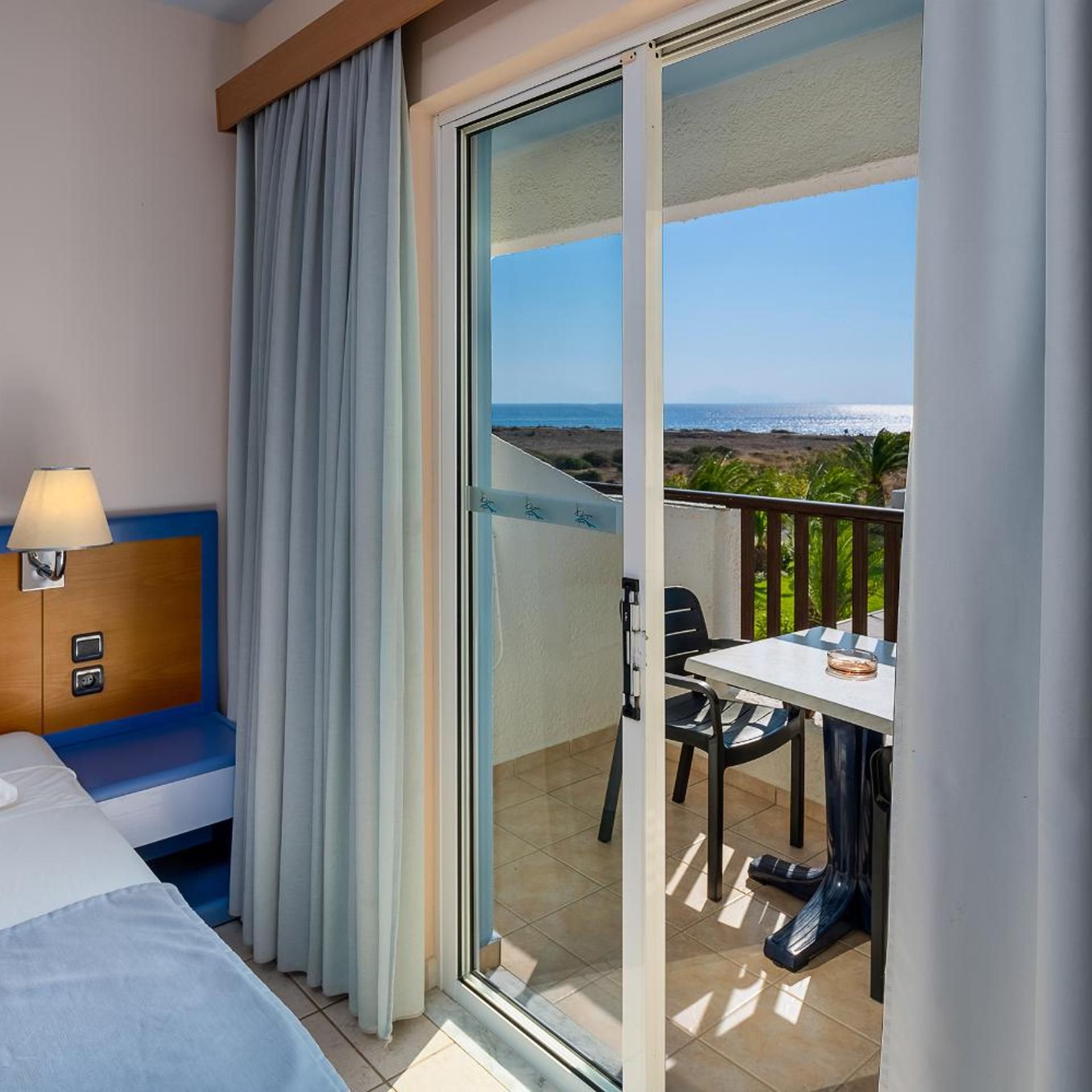 A hotel room with a bed, a desk with a lamp, a chair, and a balcony with a table and a view of the beach.