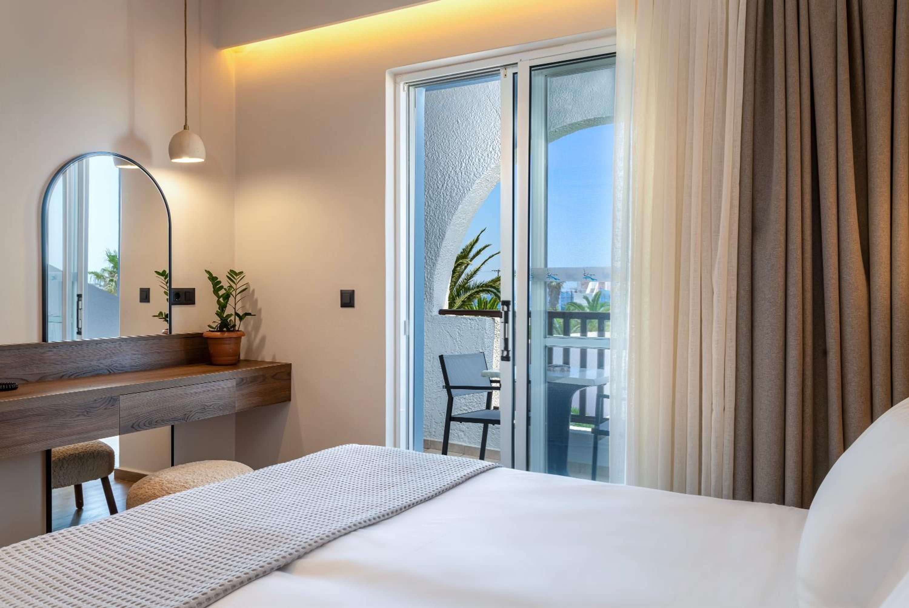 Ein Hotelzimmer mit einem Bett, einem Schreibtisch mit Stuhl, einer Topfpflanze, einem Spiegel und einer Schiebetür zu einem Außenbereich.