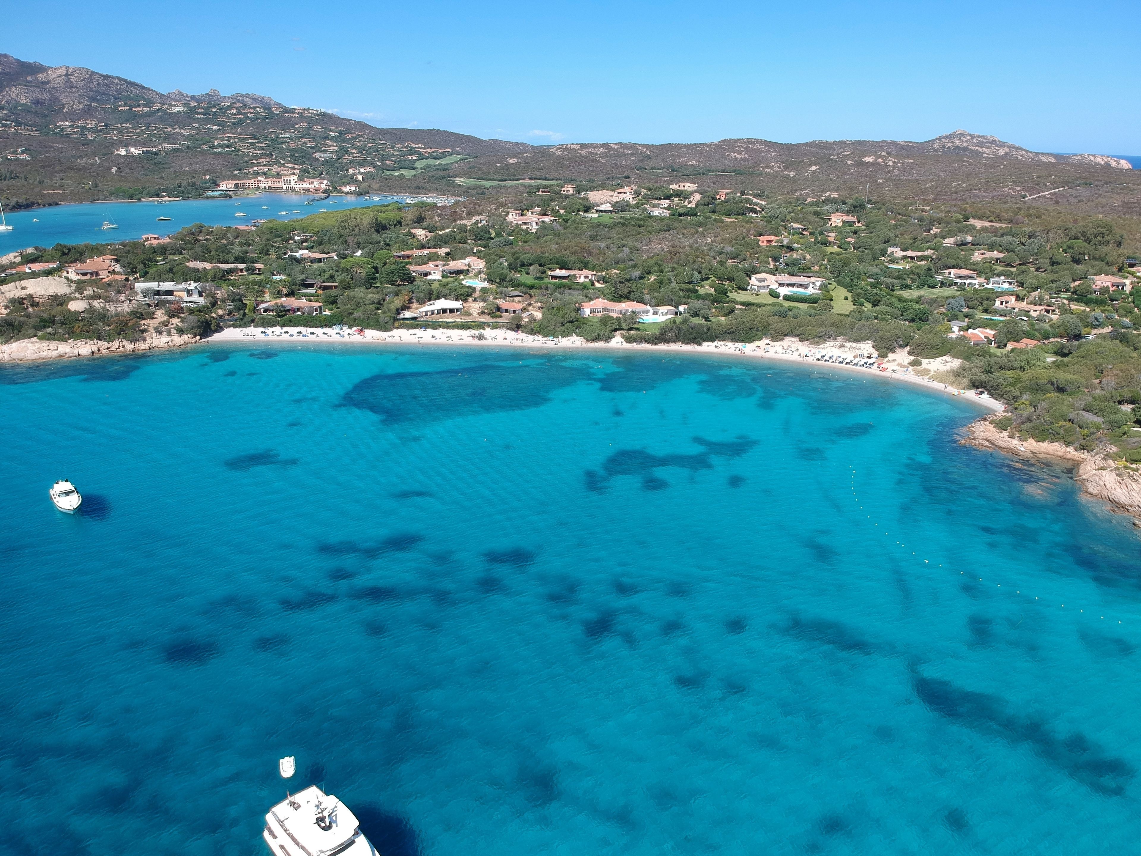 Vue aérienne d’une zone côtière avec des eaux d’un bleu limpide, un bateau et un paysage vallonné en arrière-plan.