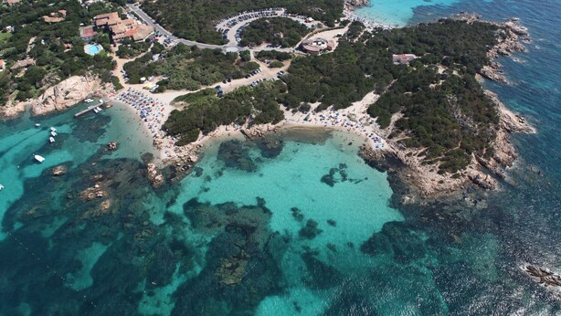 An aerial view of a coastal area with a beach, parking lot, and buildings. - Residenza Capriccioli (Arzachena)