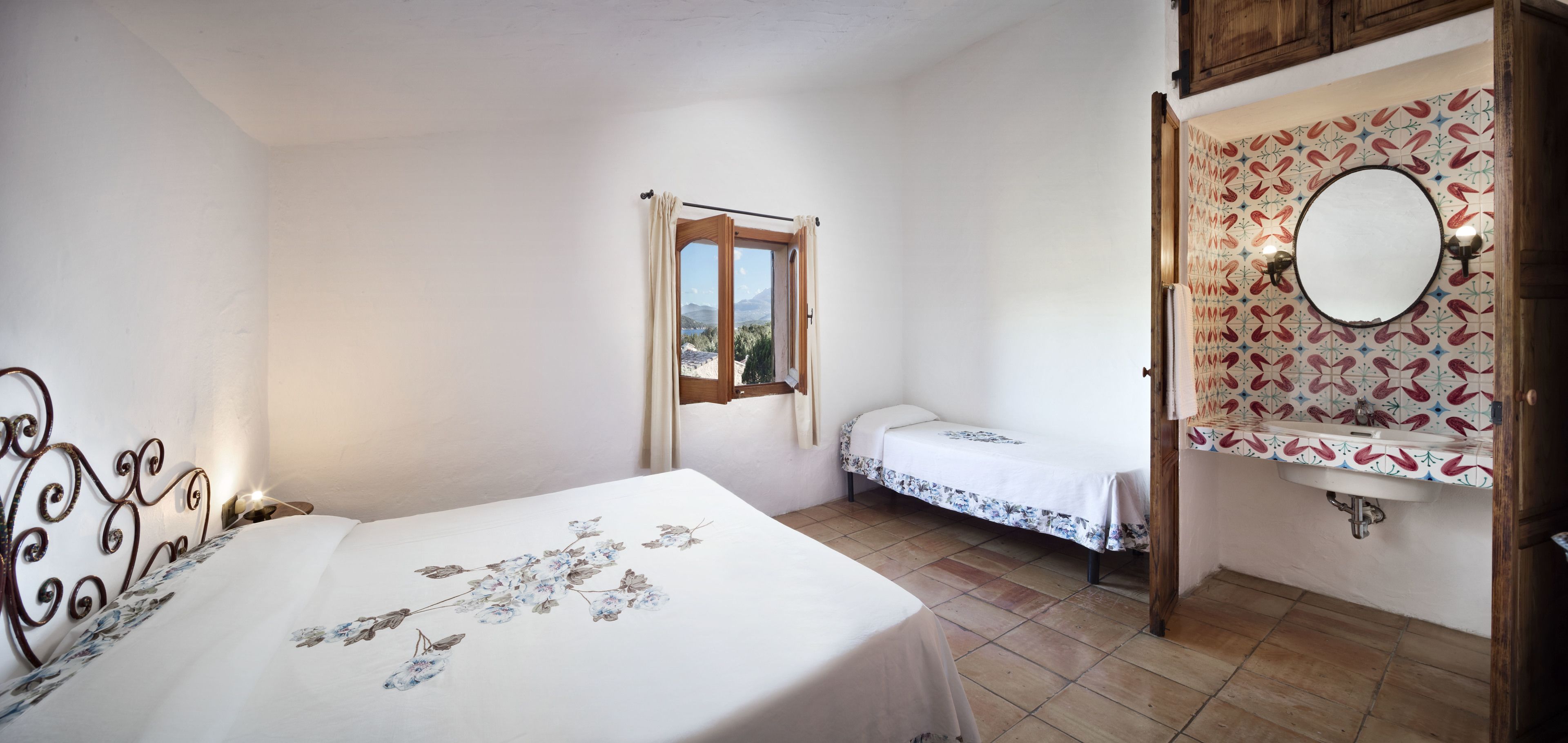 A bedroom with a bed, a window, a bedside table, a nightstand, a mirror, and a bathroom with a sink and patterned tiles.