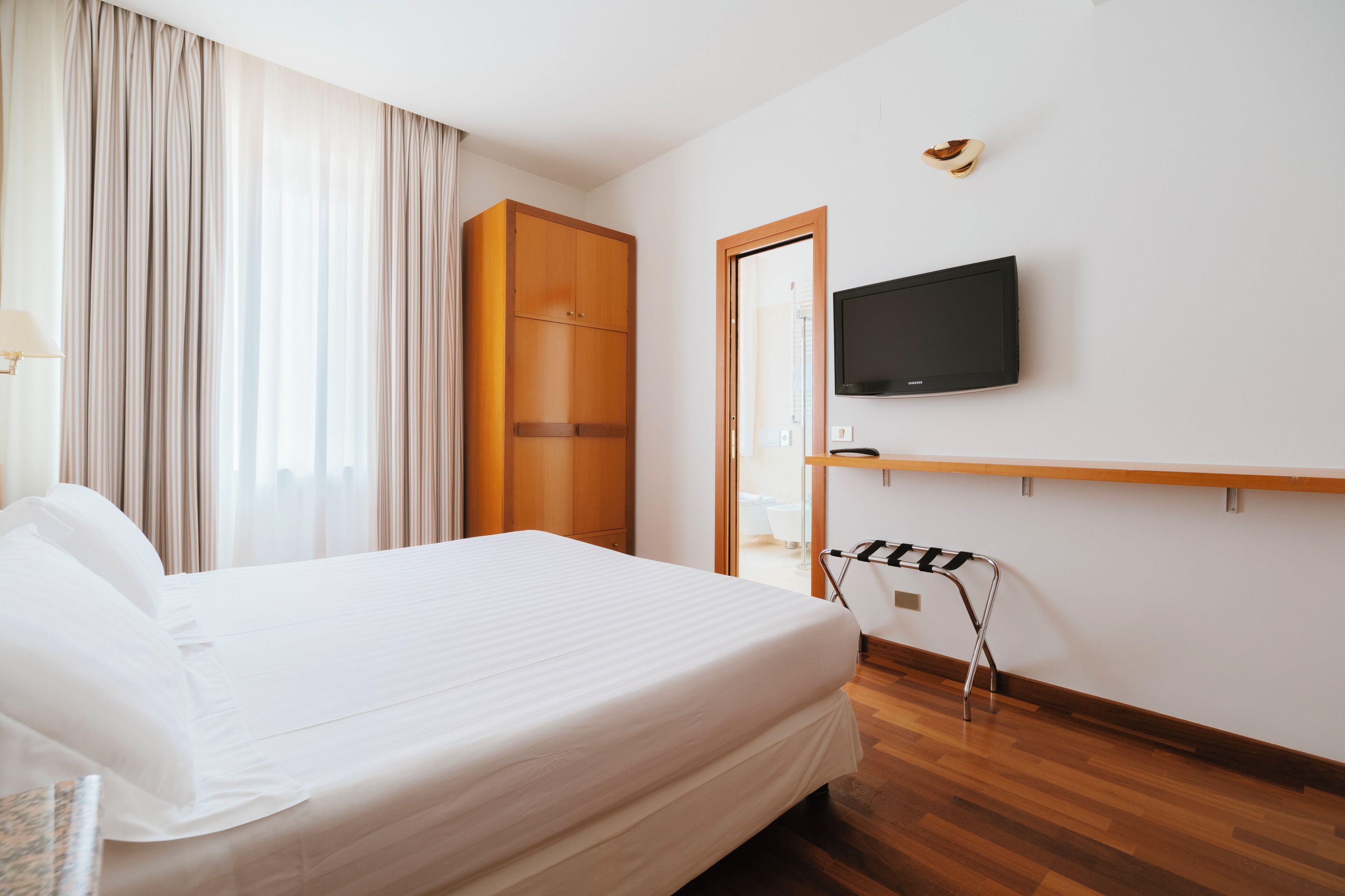A hotel room with a bed, a television mounted on the wall, a wooden wardrobe, and a wooden floor.