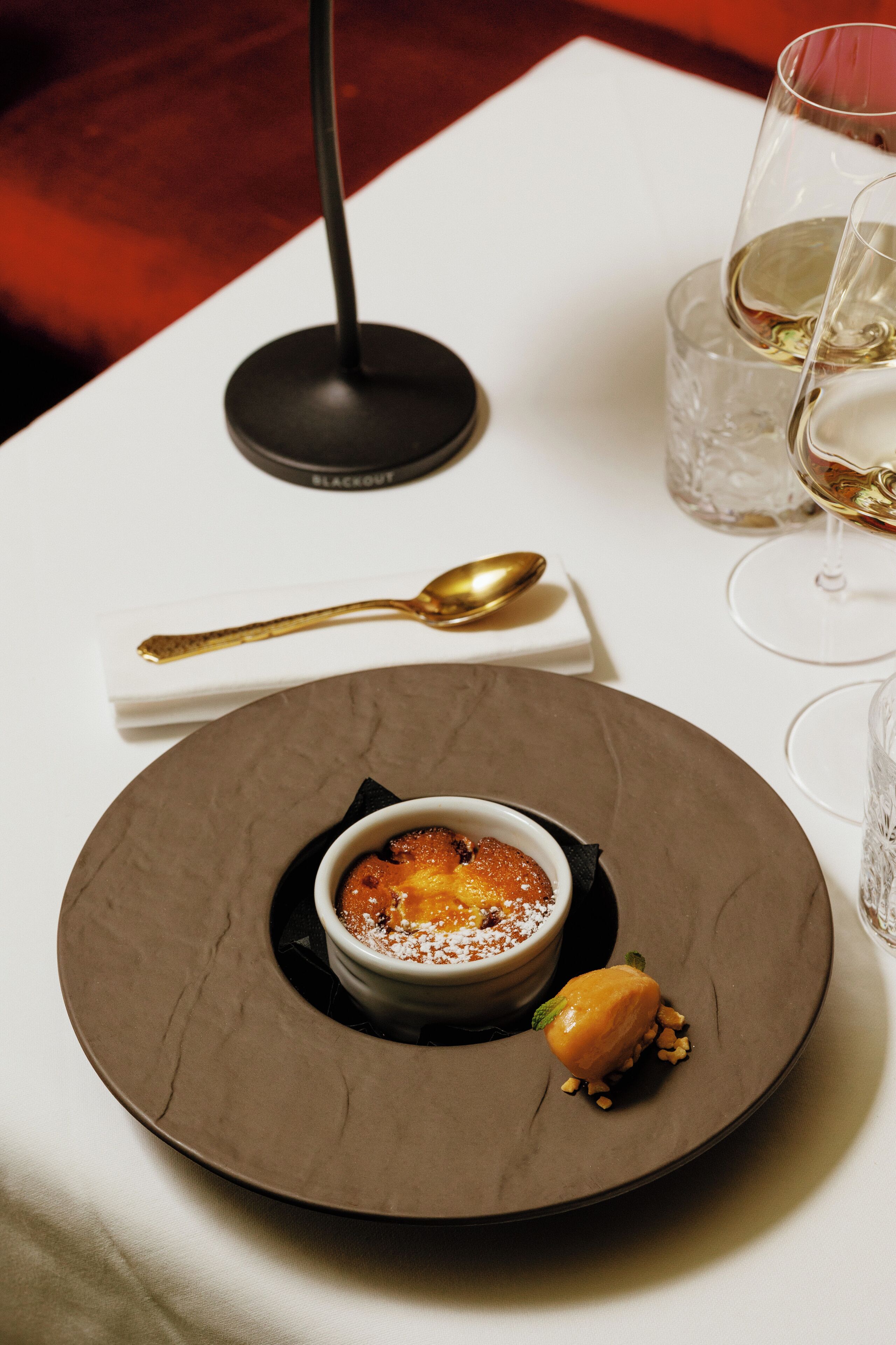 A dessert on a plate with a spoon and a glass of white wine on the table.