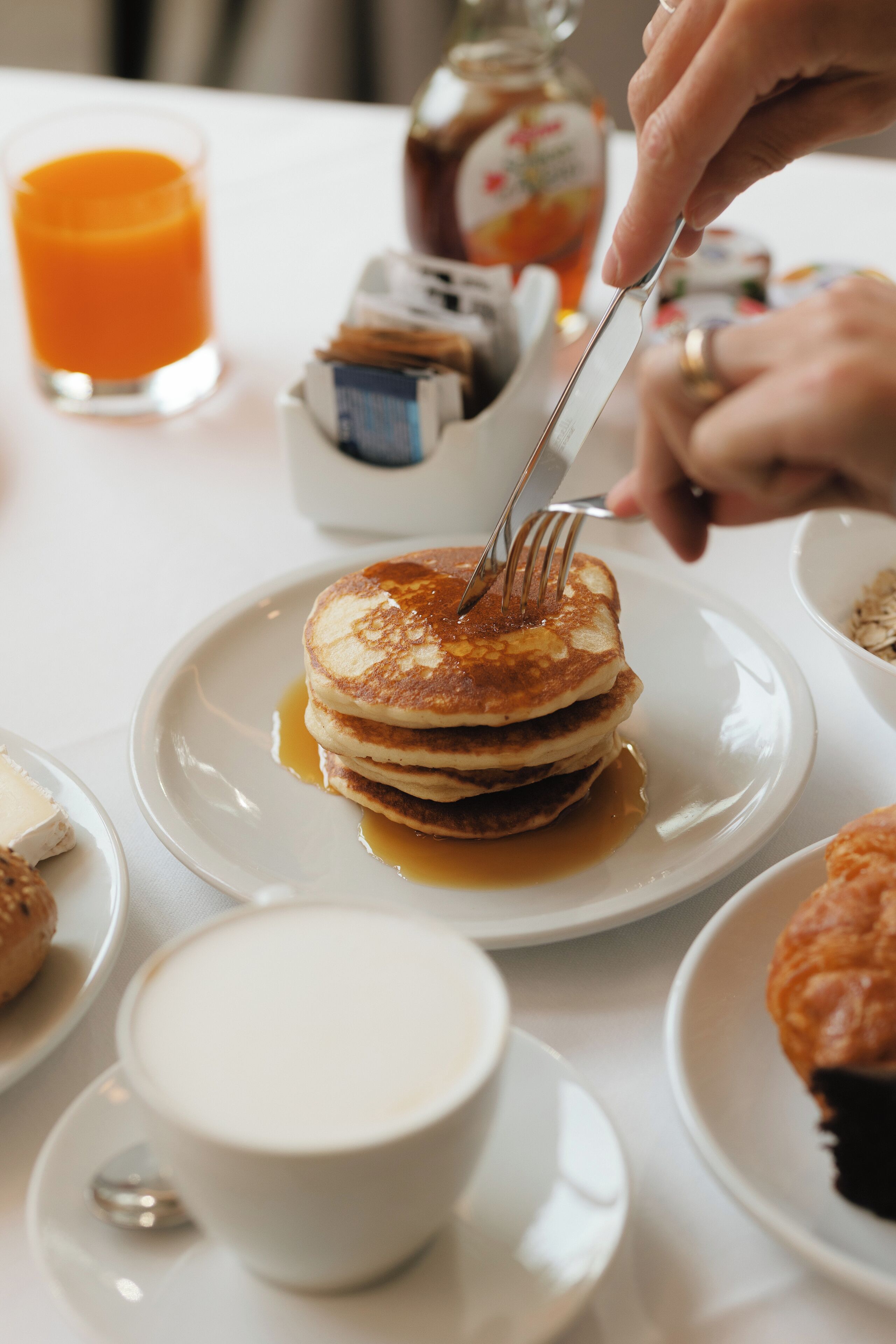 A breakfast setting with pancakes, syrup, a glass of orange juice, and a jar of jam.