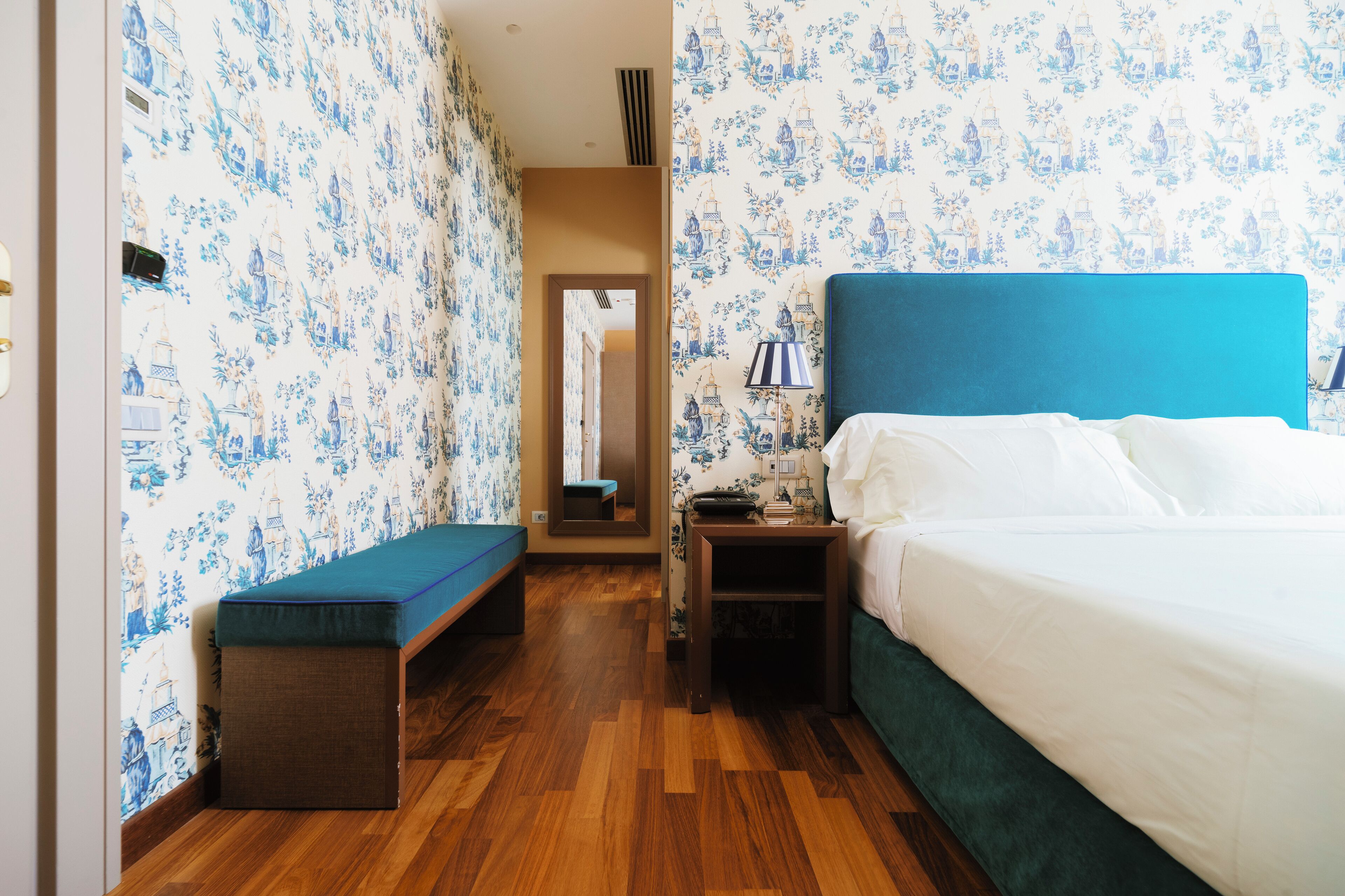 A hotel room with a bed, a bench, a nightstand, and a mirror.