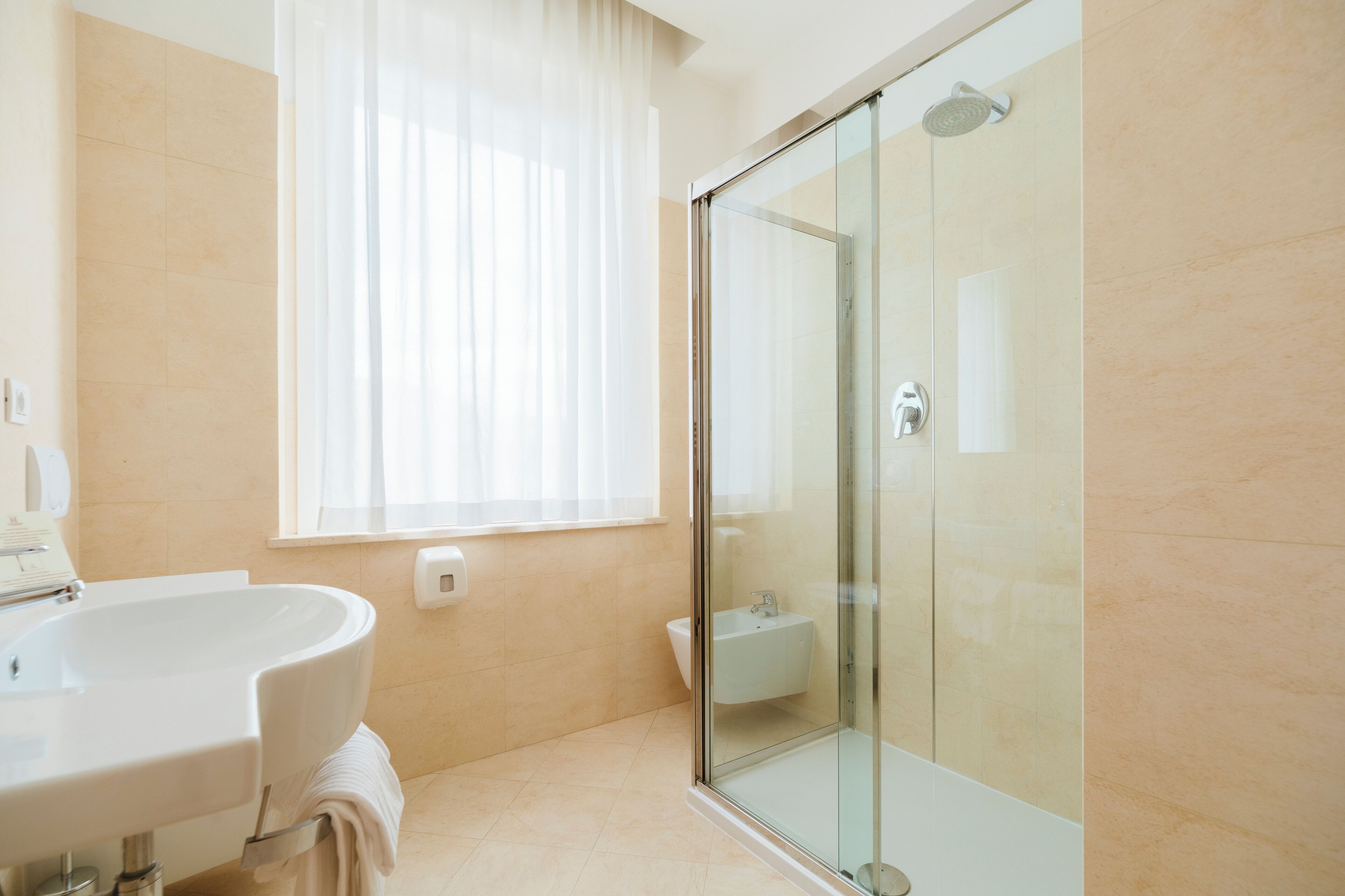 Un baño moderno con cabina de ducha de vidrio, un lavamanos y una ventana con cortinas translúcidas.