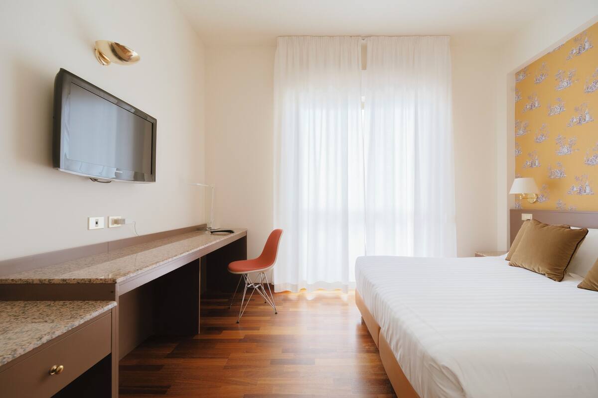 A hotel room with a bed, a desk with a chair, a television, and a window with curtains.