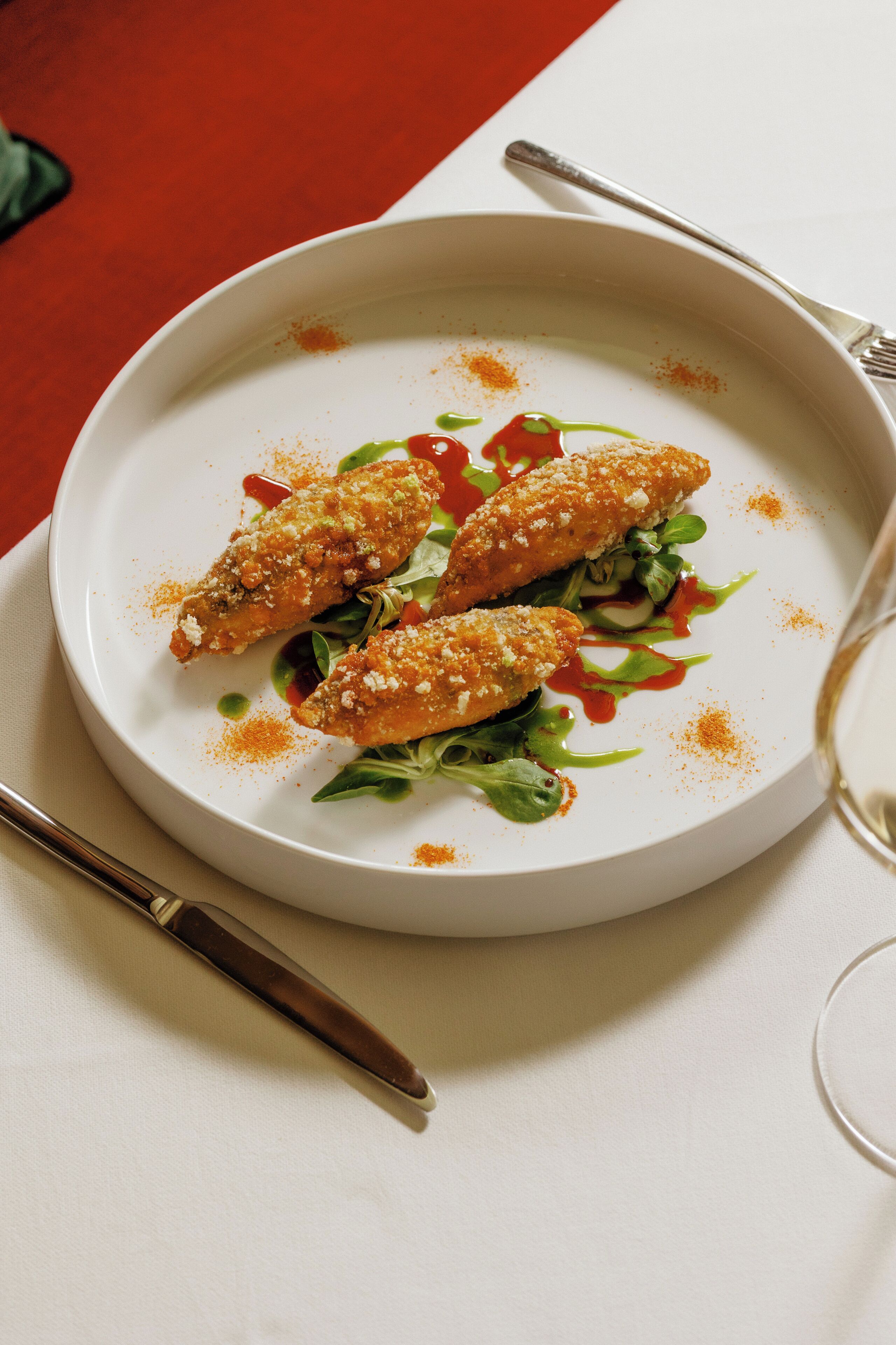 Un plato blanco con dos porciones empanizadas, vegetales verdes y salsa roja sobre una mesa con tenedor y cuchillo.